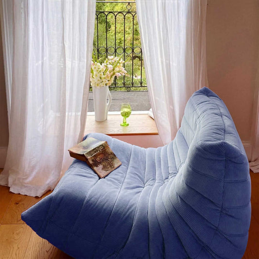 Blue corduroy Togo armchair with quilting detail placed near a window with sheer curtains and a wooden floor.