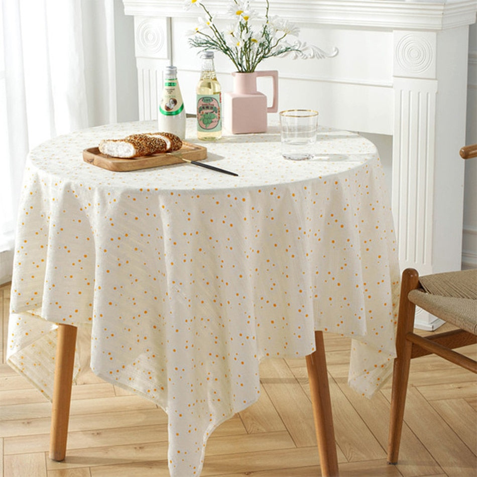 White cotton tablecloth with floral daisy pattern and tassels on a round coffee table in a bright room.