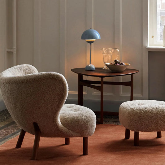 Little Petra Lounge Chair with ottoman in cozy faux sheepskin fabric and cherry wood legs next to a wooden side table with lamp in a stylish living room setting