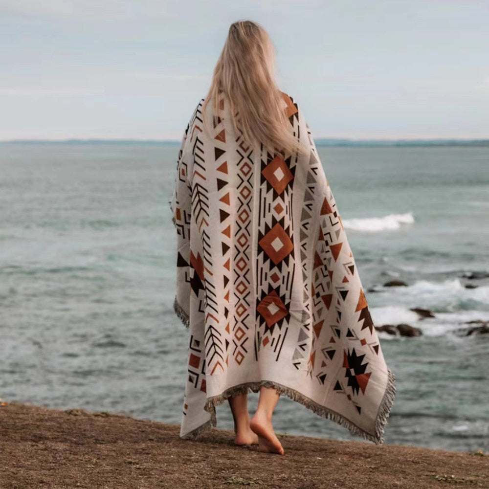 Person standing by the ocean wrapped in a versatile, knitted beach blanket with a yarn-dyed pattern.