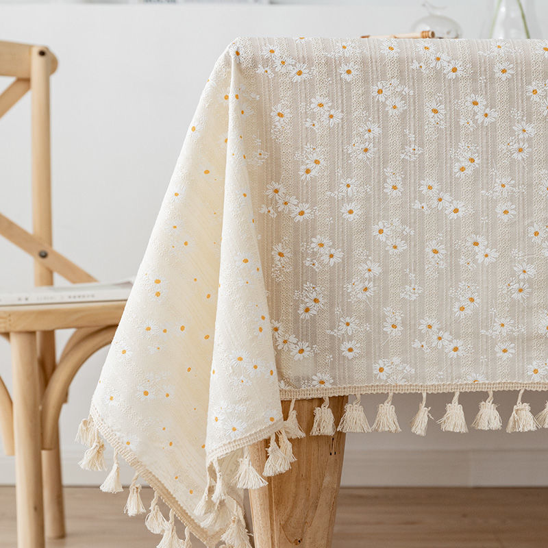 White tablecloth with floral daisy pattern and tassels on a wooden table.