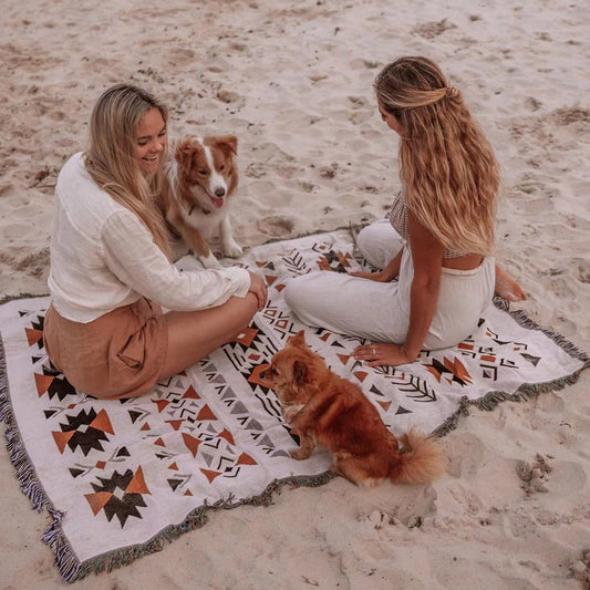 Linen and cotton beach blanket on sand with two women and two dogs.