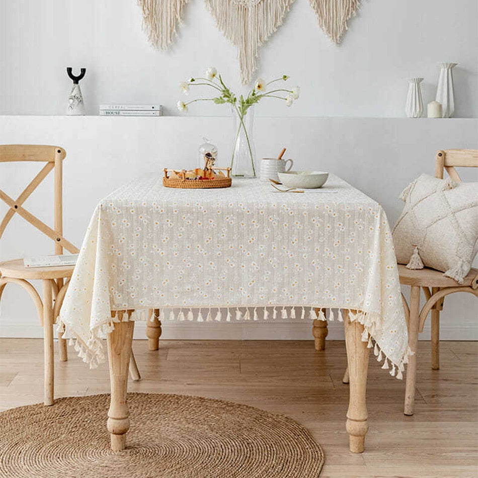 White tablecloth with floral daisy pattern and tassels on wooden dining table, enhancing elegant decor.